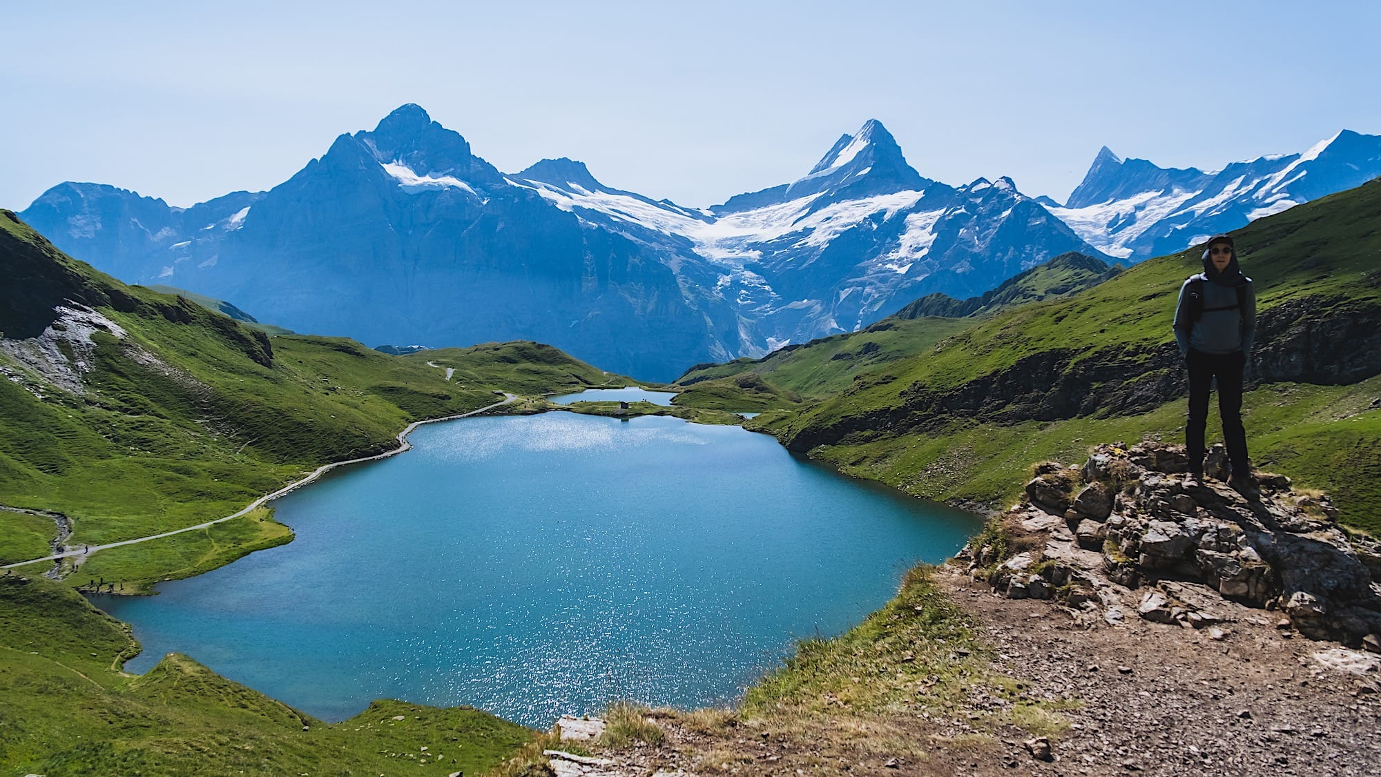 Elevated view of both Bachalpsee lakes with a hiker on the rocks and jagged peaks behind