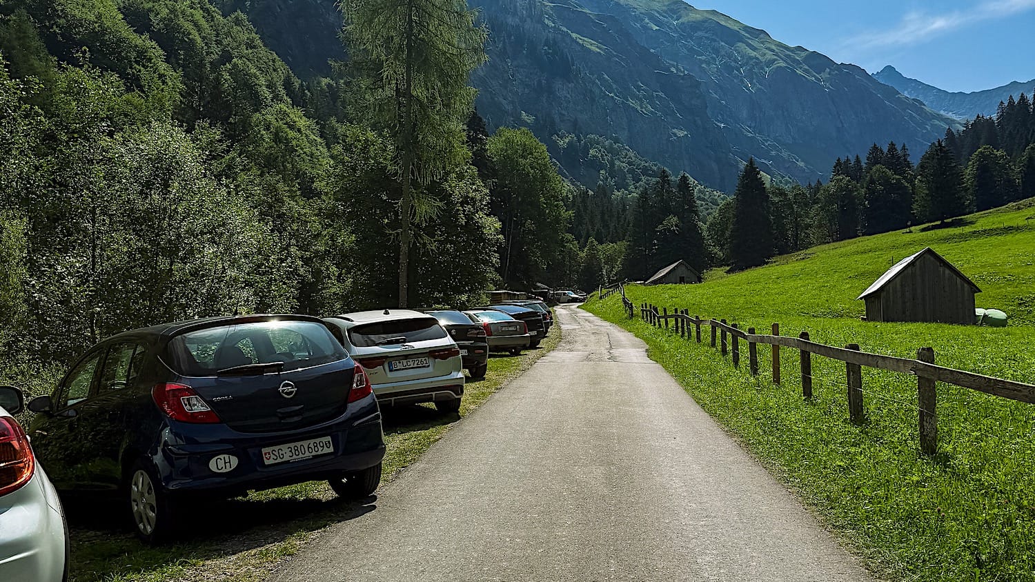 Cars parked along the road at the start of the Batöni trail near Weisstannen, with a mountain meadow ahead
