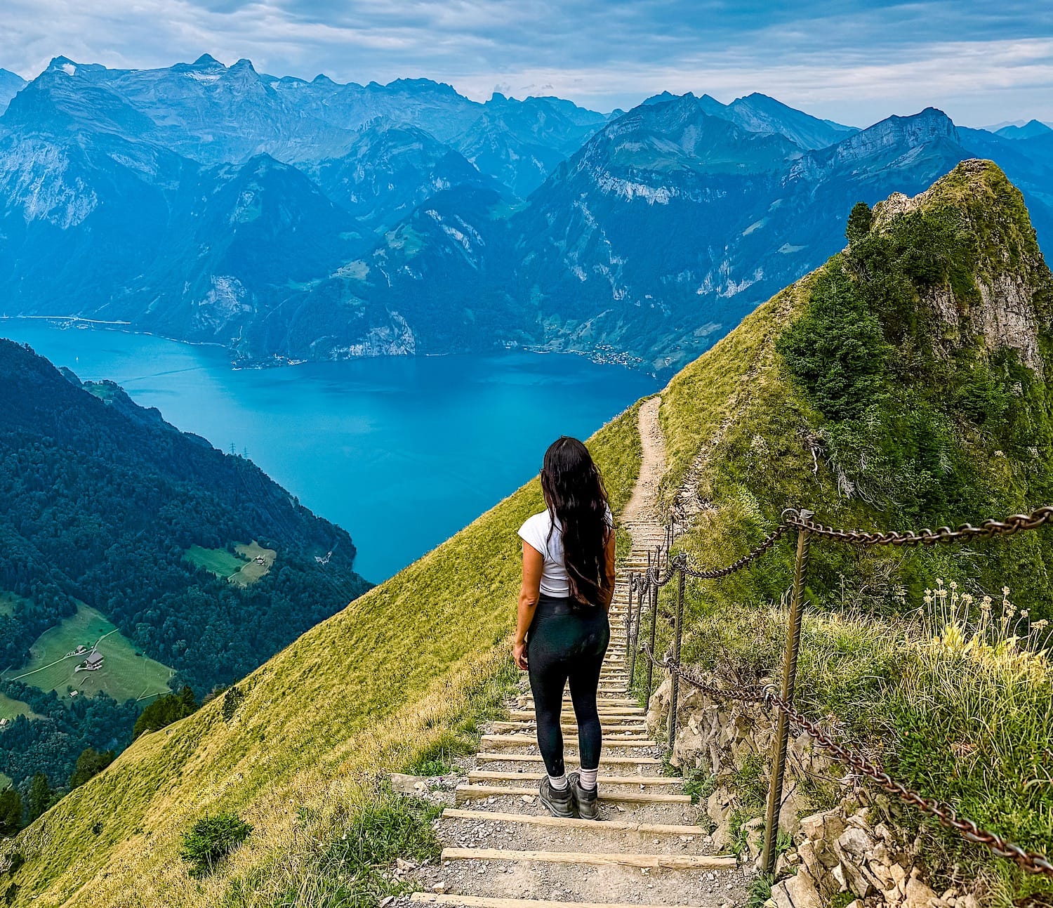 Woman standing on a trail made out of stairs, next to a green hill, with a blue lake in the background surrounded by dramatic mountains