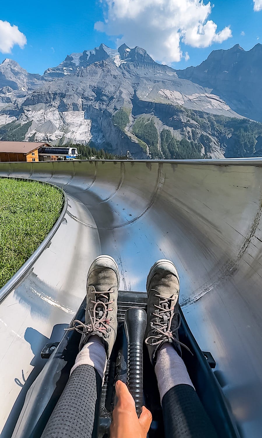 The feet of a person sitting on a sleight in a metal toboggan. A hand is holding the brake. Mountains on the background