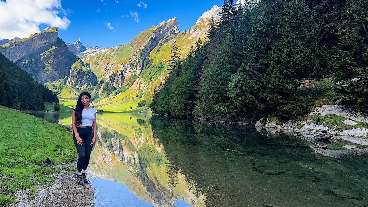 Woman standing on the shore of Seealpsee, mountains and forest mirrored in the still water