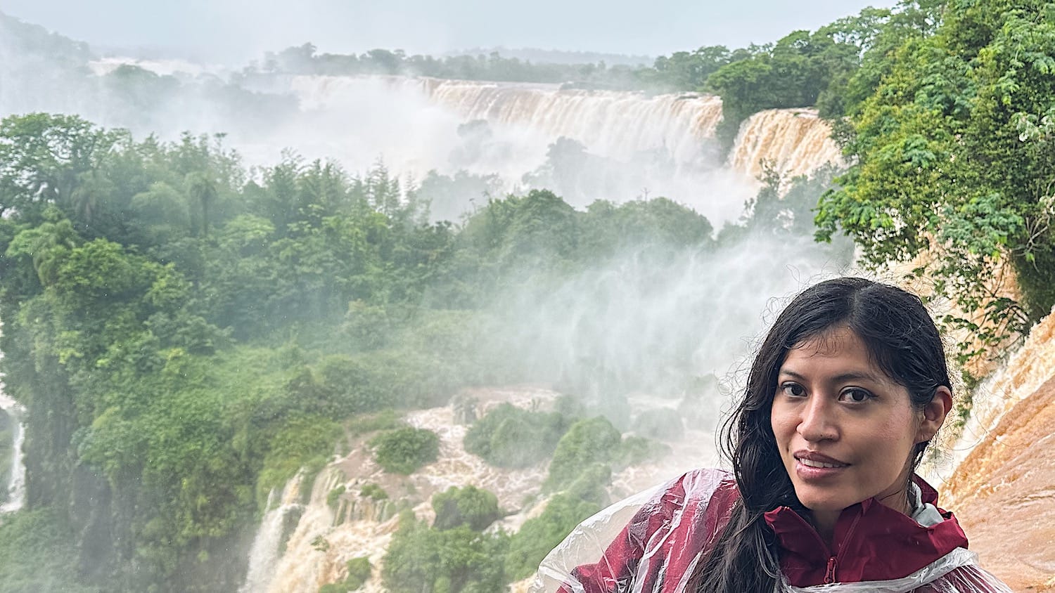 Woman in a red rain poncho in front of Iguazu Falls, with mist rising over the waterfalls and rainforest on an overcast day