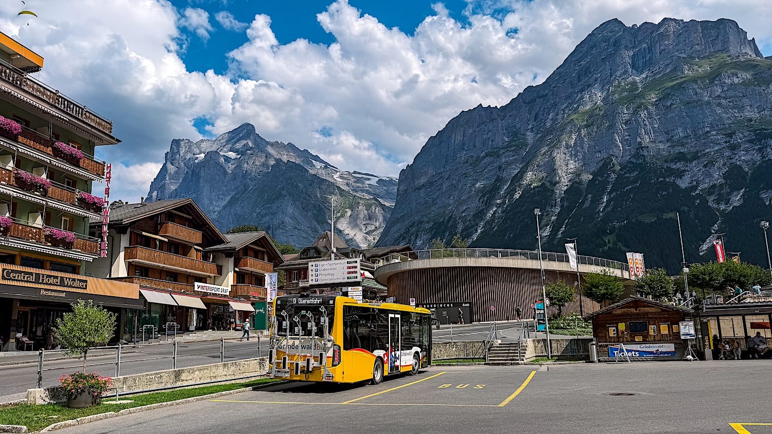 Yellow bus on the road to the Pfingstegg cable car with chalets and mountains behind