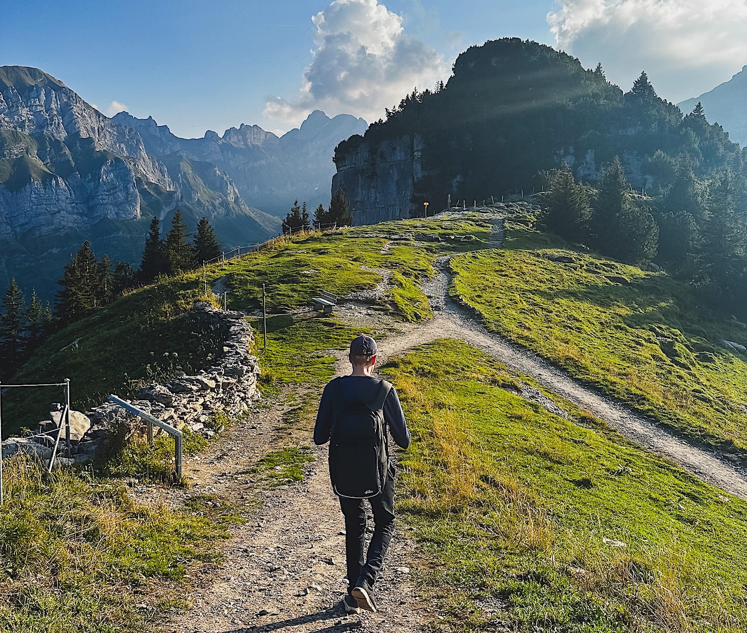 Hiker with a backpack on a gravel path along a green ridge, Alpstein peaks to the left