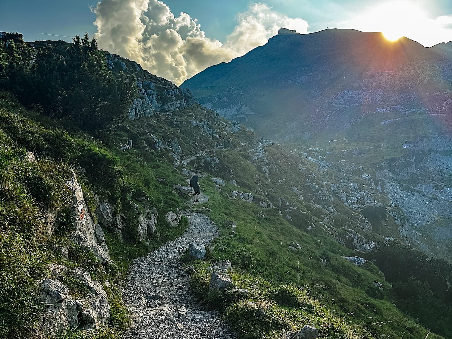 Rocky trail traversing a steep slope at sunset, a hiker ahead and a mountain hut on the ridge above