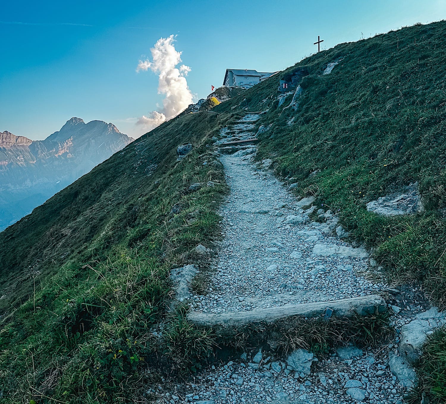 Stepped gravel path climbing to Schäfler Hut, with a summit cross and Swiss flag at the top