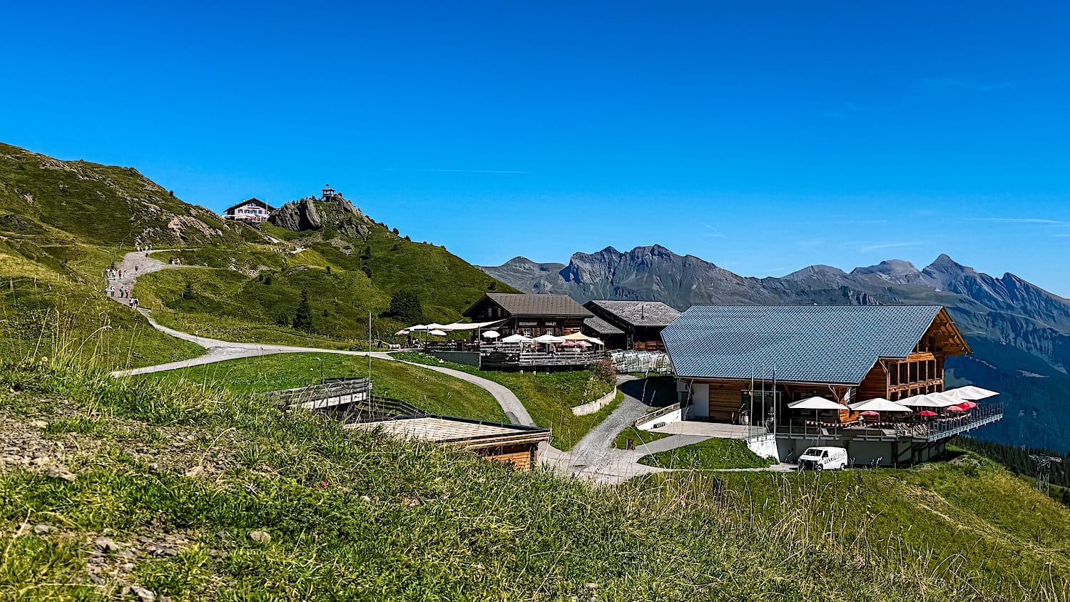 Mountain restaurant and chalets on a green alpine slope with a trail winding past under a blue sky