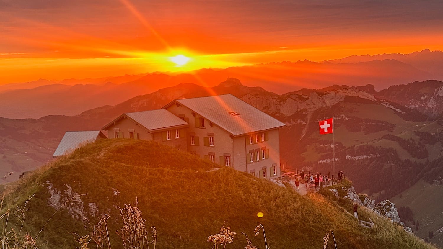 Schäfler Hut at sunrise under an orange sky, Swiss flag flying and guests on the terrace