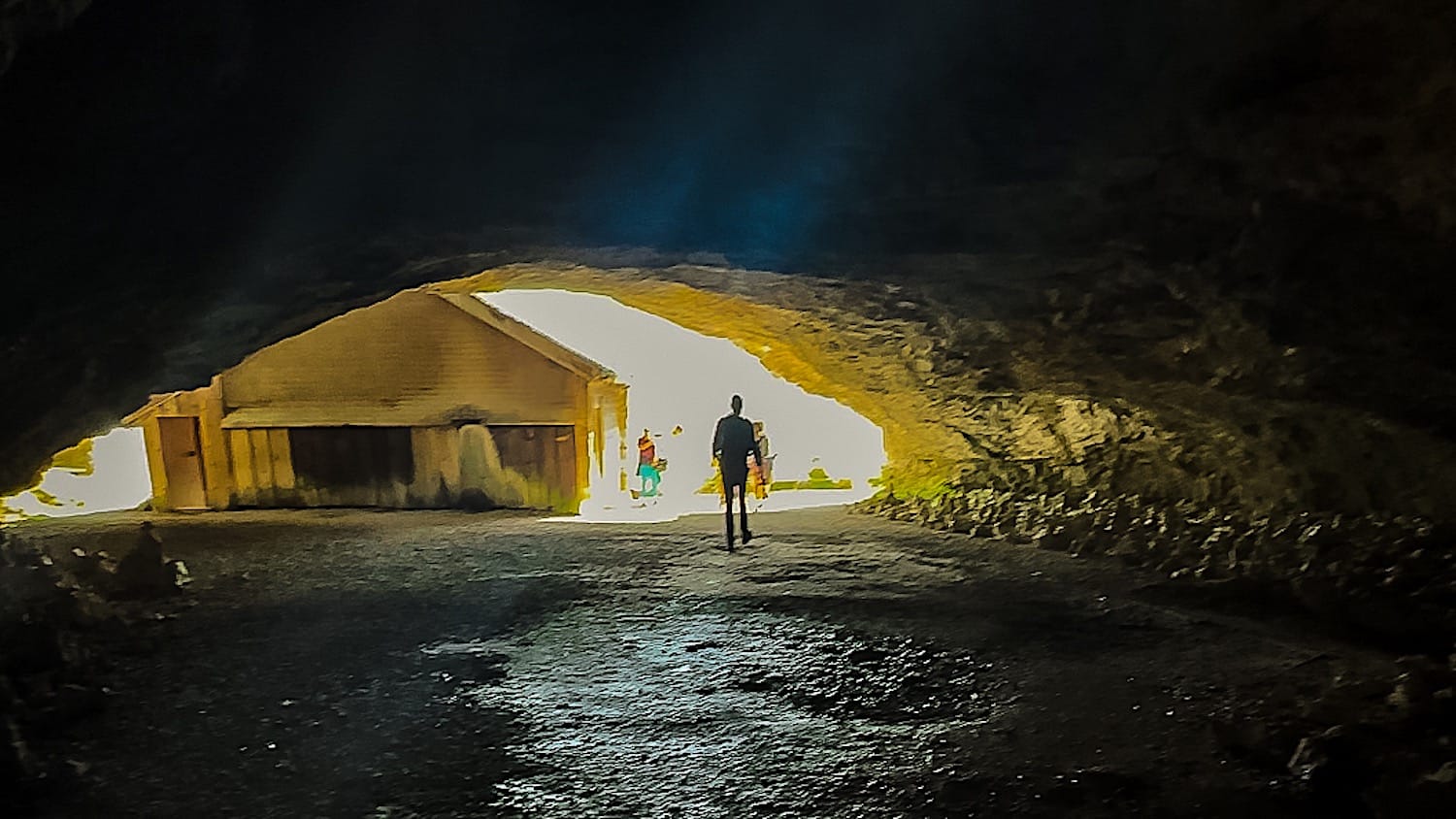 Hiker walking through the dark cave toward the bright opening, a wooden shed inside