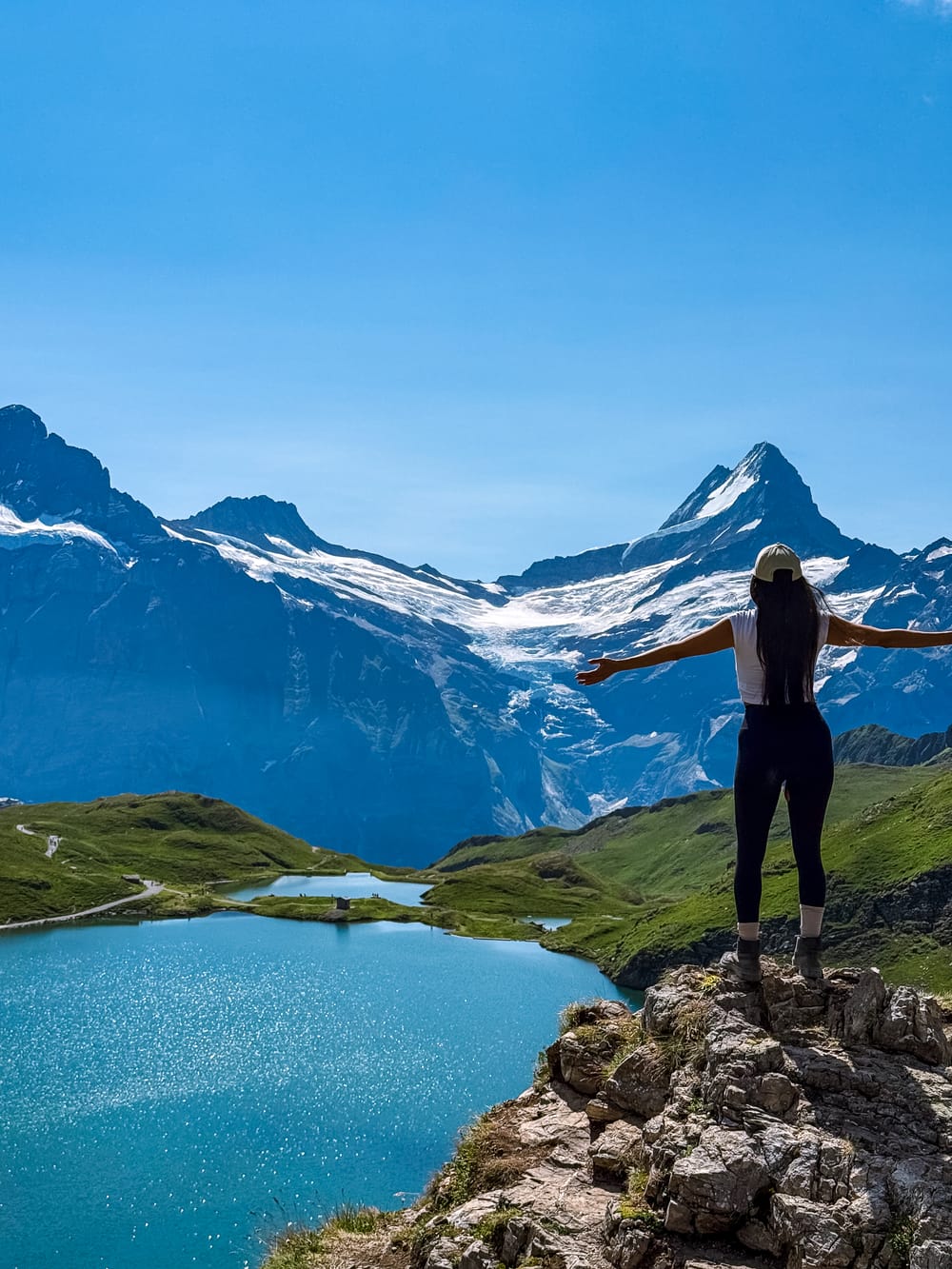 Elevated view of both Bachalpsee lakes with female a hiker on the rocks and jagged peaks behind, arms open